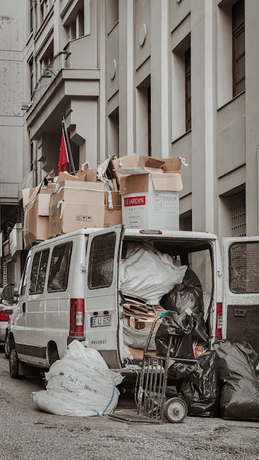 A white cargo van equipped for house relocation parked on a city street in Stepney, with its rear doors open revealing various packed moving materials. Inside the van, large black plastic waste bags, cardboard boxes, and wooden furniture parts are visible, ready for transportation. Several cardboard boxes, some sealed with packing tape and others open with packaging materials visible, are stacked on top of the van and on the pavement nearby. A metal trolley loaded with smaller boxes and items is positioned next to the open rear of the van, indicating a loading or unloading process. The surrounding environment includes multi-storey residential buildings with beige facades and windows, and the lighting appears natural, suggesting daytime. The scene captures elements of home removal logistics, with furniture, packing materials, and equipment associated with professional removals, as managed by Man With a Van Stepney and their removal services.