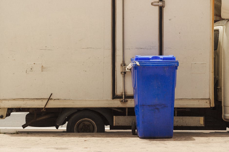 A blue plastic recycling bin is positioned on the pavement beside a white delivery van with a closed cargo compartment, which is parked close to a building. The van’s side door is open, revealing a partially visible interior, possibly used for furniture or household items. The van appears to be in the process of a home relocation or furniture transport, as indicated by the loading area and the proximity to the building entrance. The background shows the lower part of the building wall and the pavement, with the van’s rear wheel and a small section of its undercarriage visible beneath the cargo area. The environment is outdoors during daylight, with a neutral lighting setting, typical of a moving or removal service operation carried out by [COMPANY_NAME]. This scene captures the logistical aspect of packing, loading, and transporting household goods during a house removal or clearance.