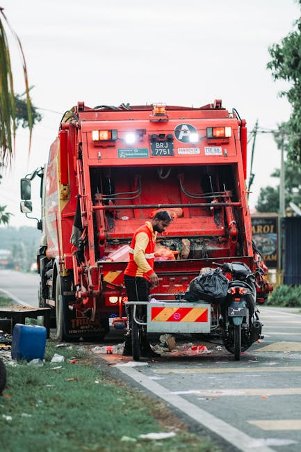 A man wearing a yellow and red high-visibility jacket operates a red waste collection vehicle with a white circular logo on the back, positioned on a street near a pavement. The rear hatch of the vehicle is open, revealing internal machinery and a loading area. The man is standing beside a black scooter parked on the pavement, which is partially loaded with black plastic bags containing waste or bulky materials such as cardboard and fabric. There are some items on the ground, including a blue container and scattered debris. The scene is outdoors during daytime, with a cloudy sky, trees in the background, and a few buildings and power lines visible. The setting suggests a waste collection or bulky waste removal activity, similar to house removals or packing logistics, consistent with the services offered by [COMPANY_NAME] at [PAGE_URL], focused on efficient furniture transport and home relocation.