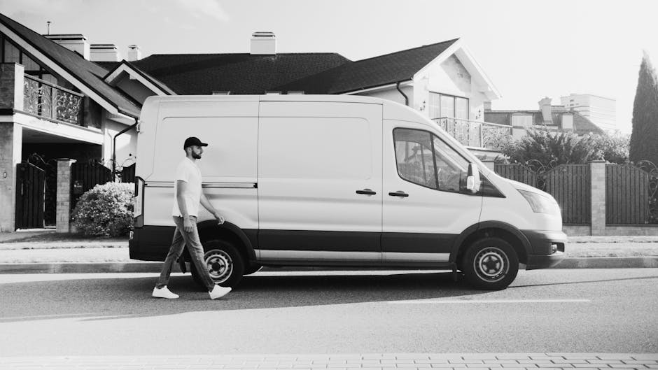 A man with a beard, wearing a white polo shirt and dark cap, stands in the open rear of a moving van loaded with numerous cardboard boxes of various sizes, some wrapped in plastic or packing materials