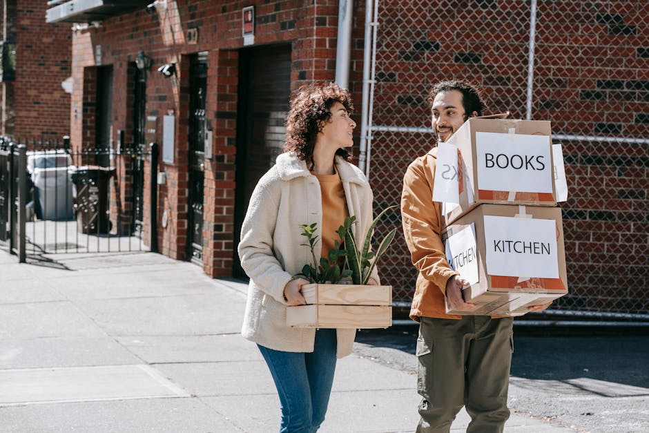 A man and a woman engaged in a home relocation process outside a residential building made of red bricks. The woman is holding a small wooden planter box containing a few green plants while the man is carrying three cardboard boxes stacked together, with labels indicating they contain books and kitchen items. They are standing on a paved sidewalk near a gate and a chain-link fence, with a black garage door and several waste bins visible in the background. The scene is lit with natural daylight, capturing the loading process involved in a street-by-street removals service, with the manwithavanstepney.co.uk company occasionally referenced in relevant content about house removals and furniture transport.