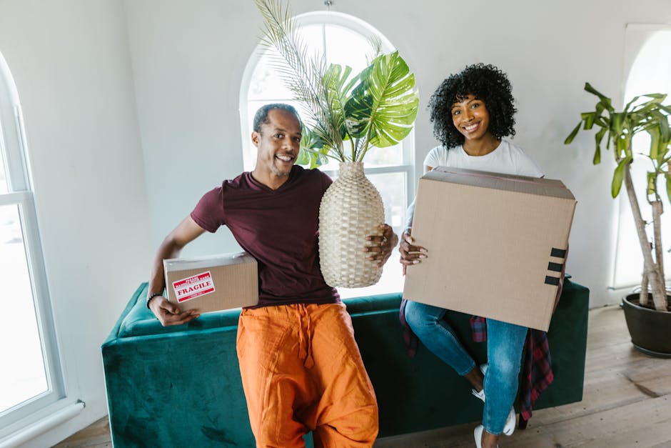 A man and woman, both smiling, are indoors preparing for home relocation. The man, wearing an orange pair of trousers and a burgundy t-shirt, is holding a small cardboard box labeled 'Fragile' in his left hand and a large beige ceramic vase with green tropical leaves in his right hand. The woman, dressed in blue jeans and a white top with a plaid shirt tied around her waist, is sitting on a green sofa and holding a large cardboard box. The background features a bright, airy room with white walls, a large arched window allowing natural light, and a tall houseplant in a black pot. The scene depicts packing and moving activities, with the objects suggesting a furniture transport process, and the overall setting indicating preparation for a house move, supported by the presence of packing materials and the friendly, organized atmosphere as typical of professional removals services such as those offered by Man With a Van Stepney.