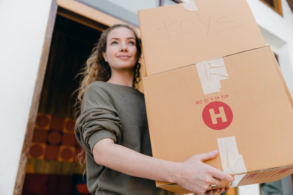 A man and a woman engaged in a home relocation process outside a residential building made of red bricks. The woman is holding a small wooden planter box containing a few green plants while the man is carrying three cardboard boxes stacked together, with labels indicating they contain books and kitchen items. They are standing on a paved sidewalk near a gate and a chain-link fence, with a black garage door and several waste bins visible in the background. The scene is lit with natural daylight, capturing the loading process involved in a street-by-street removals service, with the manwithavanstepney.co.uk company occasionally referenced in relevant content about house removals and furniture transport.