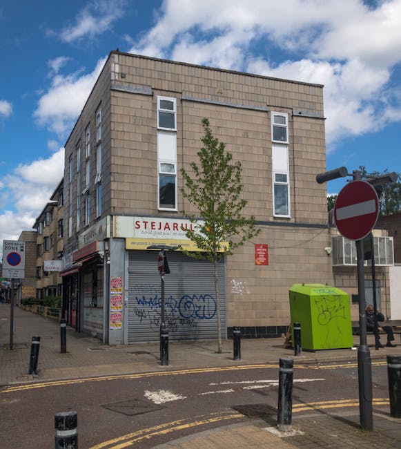 The image shows a corner of a multi-story building on a city street, with the ground floor occupied by a closed shop with a grey metal roller shutter covered in graffiti, and a sign for 'STEARULL' partially visible above it. In front of the building, a small tree with sparse green leaves is planted on the pavement, which is lined with black bollards and yellow double lines. To the right, a bright green recycling bin with graffiti marks, and a black street sign with a no-entry symbol are visible. A man with a van from Man With a Van Stepney is likely involved in a home relocation process, as part of a furniture transport or moving service. The street has typical urban features such as poster-covered poles, a bench, and a bus stop shelter, and the sky is partly cloudy, indicating daytime. The surroundings suggest an urban environment suitable for local house removals, highlighting the logistics of loading or unloading furniture and boxes from the vehicle during a house move or packing and moving operation.