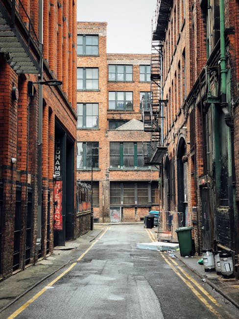 A narrow urban alleyway flanked by tall, multi-storey brick buildings with fire escapes and external staircases on both sides. The pavement is wet, with double yellow lines along the edges indicating parking restrictions. On the right side near the foreground, there are green and black wheelie bins, along with some small miscellaneous items and a parked bicycle. The alley appears to be used for service access or deliveries, with no visible pedestrians or vehicles, leading towards a larger brick building with multiple windows at the end. This setting exemplifies the typical environment in which house removals and furniture transport services, such as those provided by Man With a Van Stepney, operate during home relocation processes, often involving navigating narrow alleys to access residential buildings in the STEPNEY area.