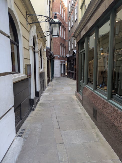 The image shows a corner of a multi-story building on a city street, with the ground floor occupied by a closed shop with a grey metal roller shutter covered in graffiti, and a sign for 'STEARULL' partially visible above it. In front of the building, a small tree with sparse green leaves is planted on the pavement, which is lined with black bollards and yellow double lines. To the right, a bright green recycling bin with graffiti marks, and a black street sign with a no-entry symbol are visible. A man with a van from Man With a Van Stepney is likely involved in a home relocation process, as part of a furniture transport or moving service. The street has typical urban features such as poster-covered poles, a bench, and a bus stop shelter, and the sky is partly cloudy, indicating daytime. The surroundings suggest an urban environment suitable for local house removals, highlighting the logistics of loading or unloading furniture and boxes from the vehicle during a house move or packing and moving operation.
