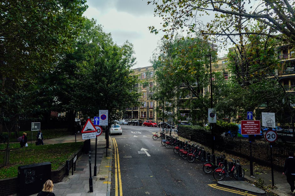 A street scene in a residential area during daytime shows a paved road with a white arrow indicating the direction of traffic flow. To the right, a row of red and black rental bikes is parked along a metal bike rack, which is situated beside a black metal fence enclosing a green space with mature trees and grass. Several street signs and parking restriction signs are mounted on posts along the sidewalk, including a no-parking zone sign and a speed limit sign of 20 miles per hour. On the left side of the street, pedestrians, including a woman with blonde hair and a pink bag, walk along the pavement in front of a small park area with leafy trees. In the background, multi-storey apartment buildings with balconies are visible, partially obscured by the trees. A white car is parked on the street, with another vehicle visible further down the road. The scene appears to be part of a home relocation or furniture transport operation, with a focus on the urban environment and traffic management related to moving services offered by Man With a Van Stepney.