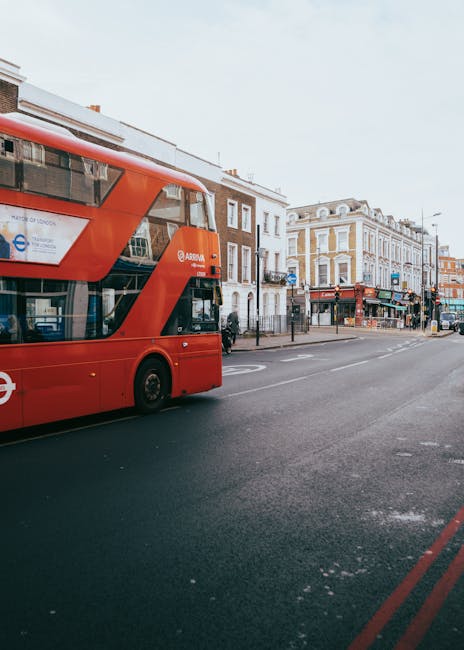 A red double-decker bus is parked beside a curb on a city street, with several multi-story buildings in the background featuring retail outlets on the ground floor. The buildings have a mix of brick and stucco facades, with some windows and decorative architectural details. The street is relatively quiet with minimal traffic, and the pavement is clean with visible road markings. The scene is captured during daylight hours, and natural light illuminates the area, highlighting the urban environment. This setting, associated with busy urban areas, is relevant to house removals and moving services offered by Man With a Van Stepney, who may facilitate furniture transport and home relocation logistics along such routes.