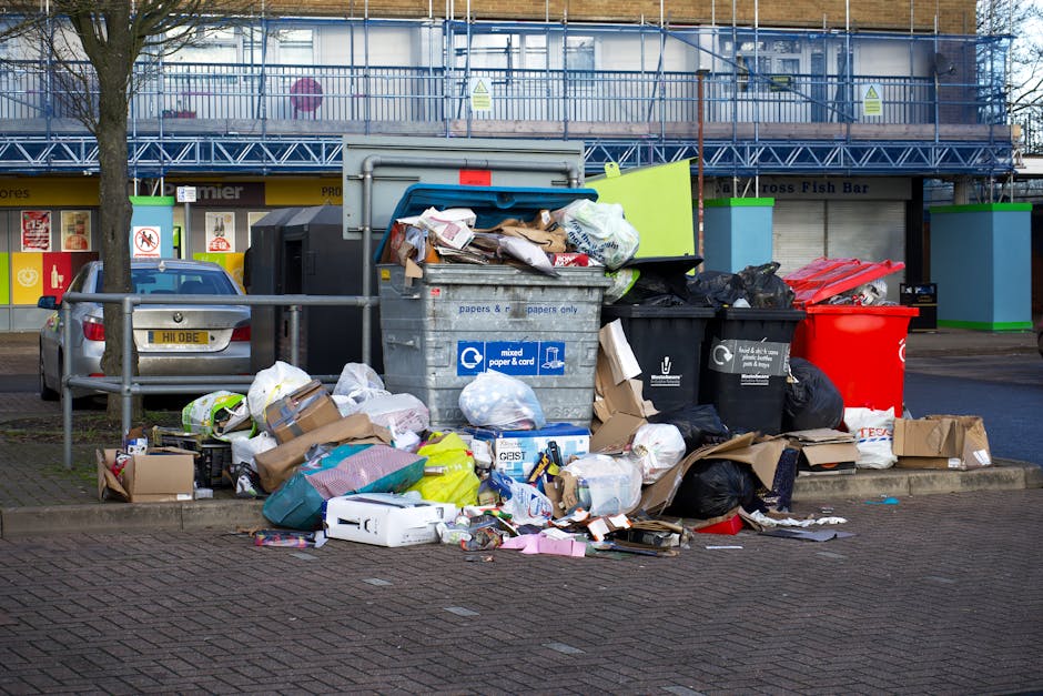 A large collection of waste and recycling bins on a paved street in front of a commercial building in Stepney, Tower Hamlets. The bins include various types such as mixed paper, general waste, and recycling containers, with open lids revealing trash inside and trash bags spilling onto the pavement. Surrounding the bins are scattered cardboard boxes, paper packaging, plastic bags, and miscellaneous waste materials. There is a parked silver car nearby, partially obstructed by a metal railing. The background features a building under construction with scaffolding, and the scene is illuminated by natural daylight, indicating daytime. This setting illustrates the importance of proper waste disposal in relation to home relocation or moving services, as handled by companies like Man With a Van Stepney, which often coordinate with waste management during packing and moving processes.