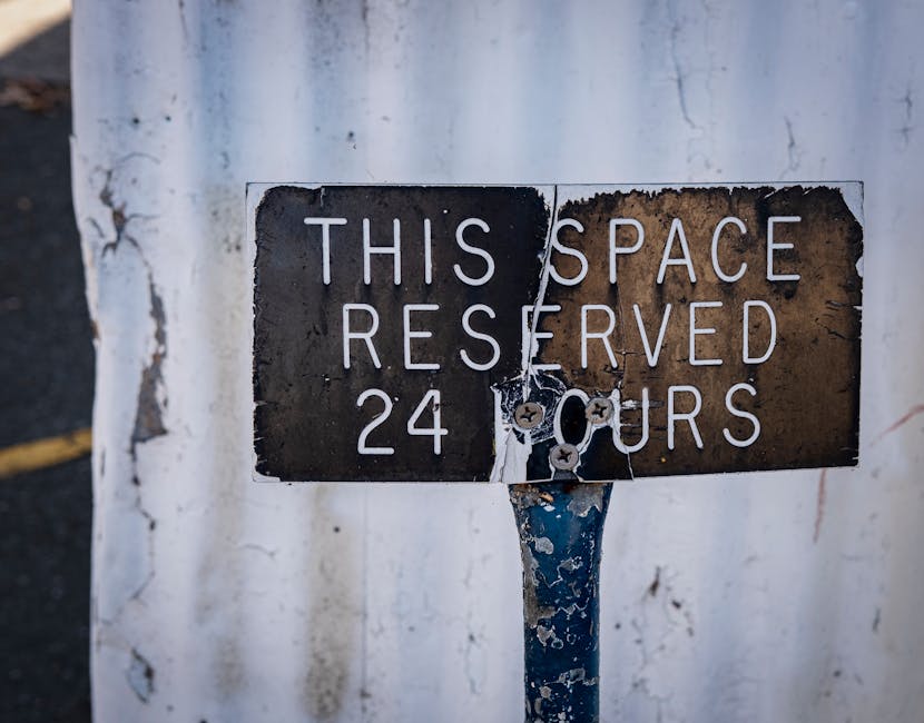 A weathered metal sign mounted on a blue pole indicating a designated parking or loading area, with the text 'This Space Reserved 24 Hours' partly obscured by torn adhesive remnants. The sign is situated against a white, slightly chipped and stained wall. In the background, a portion of a dark road or pavement is visible, suggesting an outdoor urban setting consistent with building access points used during home relocation or furniture transport. The sign's condition and placement imply restrictions on parking or loading, relevant to moving logistics and clearance processes managed by Man With a Van Stepney, as part of their removals and relocation services.