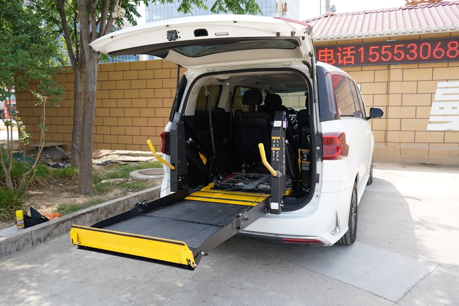 A white vehicle with its rear hatch open, showing a wheelchair accessible lift platform extended from the back of the van onto the pavement. The lift is black with yellow safety edges and features handrails on either side. Inside the vehicle, the interior is visible with black seats and equipment, and the lift is positioned to facilitate the loading or unloading of mobility aids or large objects during a home relocation or furniture transport process. The setting includes an outdoor area with a beige brick wall, trees, and a red-tiled roof structure nearby, indicating a residential or commercial environment. The image captures a professional move by [COMPANY_NAME], specialized in removals and logistics, emphasizing accessible transport solutions and proper handling of larger items during packing and moving tasks.