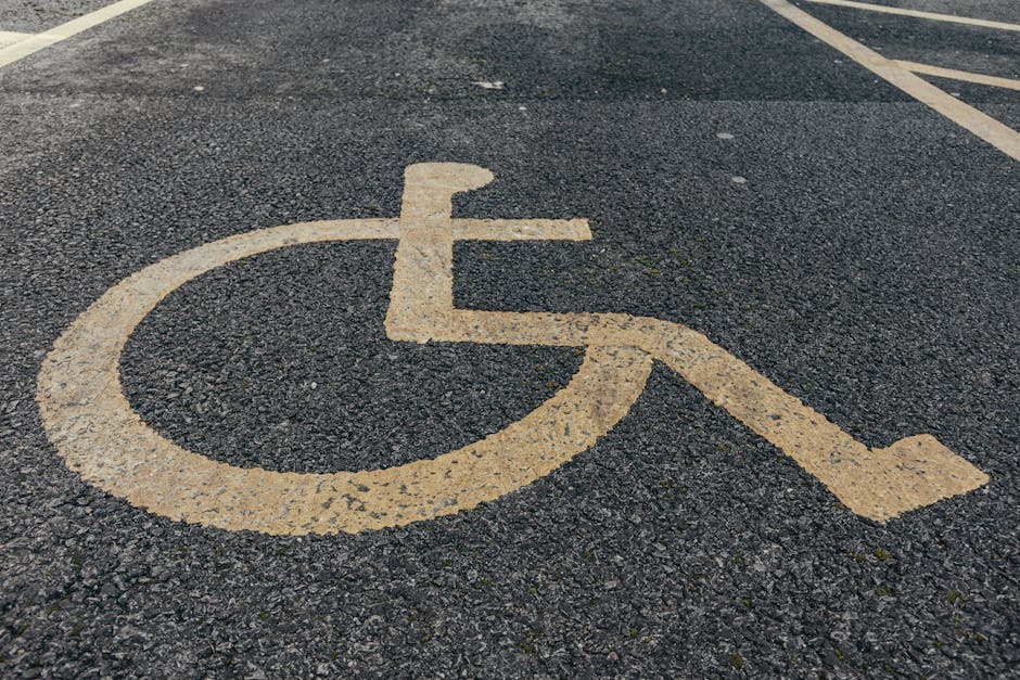 A close-up view of a designated accessible parking space on an asphalt surface, featuring a large, yellow wheelchair symbol painted on the ground with visible wear and textured detail. The parking space is part of a broader parking lot with faint white lines indicating multiple spaces. The surface appears rough with small stones embedded in the black asphalt. This image illustrates the area where vehicles engaged in home relocation or furniture transport services, such as those provided by Man With a Van Stepney, may load or unload belongings during a move. The wheelchair symbol signifies accessibility for mobility aids, and the surrounding environment suggests an outdoor parking zone suitable for a moving vehicle, with natural lighting and shadows cast by the sun. Such designated areas are important for efficient and accessible moving logistics, supporting safe and convenient service execution for house removals and packing and moving processes near Stepney Green Park.