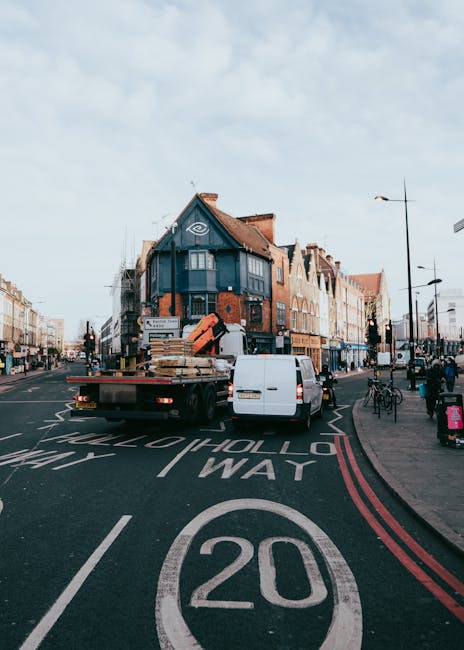 A white flatbed truck belonging to Man With a Van Stepney is parked in the middle of a busy street in Tower Hamlets, London, during the daytime. The truck is loaded with various furniture items, including wooden pallets, cardboard boxes, and wrapped household goods, indicating a home relocation or moving process. The truck's cargo area is partially covered with protective materials such as blankets and plastic wrap, and a small white van is parked adjacent to it. In the background, there are multi-storey brick buildings with retail shops, some with signage, and several pedestrians walking along the pavement. Bicycles are parked nearby, and the street features typical London road markings, including a 20 mph speed limit sign painted on the road surface. The scene captures the logistics of furniture transport and loading for house removals in an urban environment, highlighting the importance of compliance with parking and loading rules in Tower Hamlets, managed by [COMPANY_NAME].