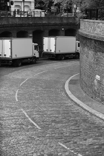 A white flatbed truck belonging to Man With a Van Stepney is parked in the middle of a busy street in Tower Hamlets, London, during the daytime. The truck is loaded with various furniture items, including wooden pallets, cardboard boxes, and wrapped household goods, indicating a home relocation or moving process. The truck's cargo area is partially covered with protective materials such as blankets and plastic wrap, and a small white van is parked adjacent to it. In the background, there are multi-storey brick buildings with retail shops, some with signage, and several pedestrians walking along the pavement. Bicycles are parked nearby, and the street features typical London road markings, including a 20 mph speed limit sign painted on the road surface. The scene captures the logistics of furniture transport and loading for house removals in an urban environment, highlighting the importance of compliance with parking and loading rules in Tower Hamlets, managed by [COMPANY_NAME].