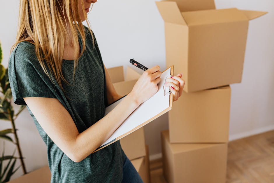 A young woman with curly hair is inside a room, sitting on a chair and writing on a clipboard or notepad, surrounded by packing materials and moving boxes. A large open cardboard box is nearby, indicating a packing and moving process as part of a home relocation. In the background, a man stands with crossed arms, observing the scene while near a window that allows natural light to illuminate the space. The room features wooden flooring and a wooden window frame, with blankets or padding possibly used to protect items during furniture transport. The overall setting appears to be an ongoing packing and moving operation, coordinated by Man With a Van Stepney, focusing on careful packing, loading, and transportation of household belongings.