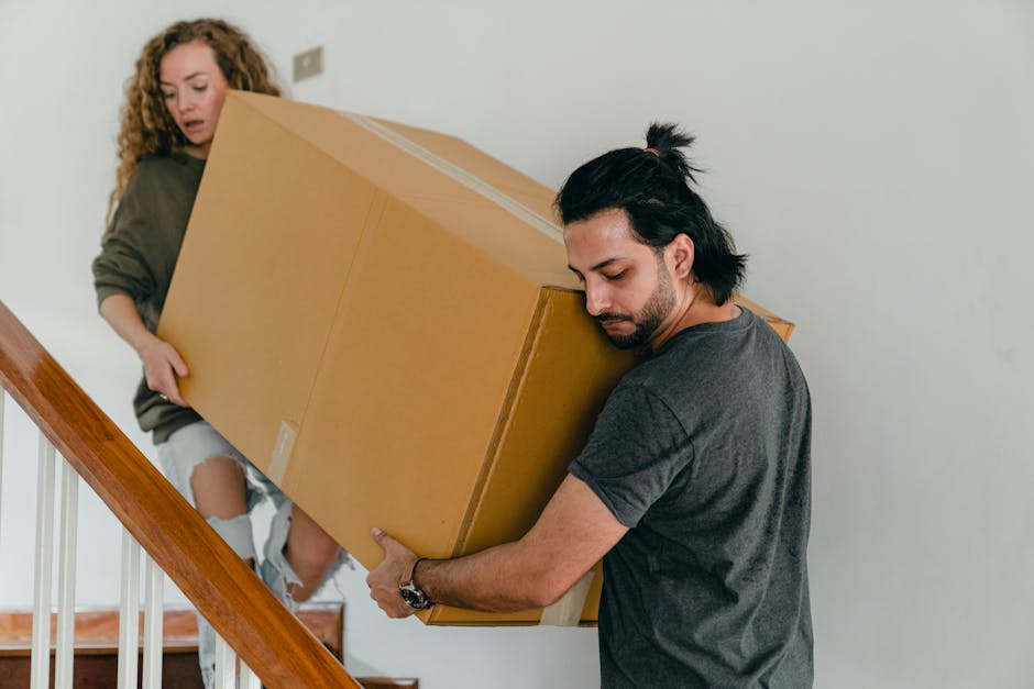 A man with dark hair tied back in a ponytail, wearing a dark grey T-shirt, is seen carefully lifting a large cardboard box while descending a wooden staircase inside a residential property. A woman with curly hair, dressed in a grey top and ripped jeans, stands nearby on the stairs, observing the process. The staircase has a wooden handrail with vertical white balusters, and the environment features plain white walls and natural indoor lighting. The large cardboard box is sealed with brown packing tape, indicating packing and moving preparations. This scene is part of a home relocation or furniture transport process, potentially managed by Man With a Van Stepney, who specialise in removals and moving services, including handling bulky items in challenging staircases or confined spaces.