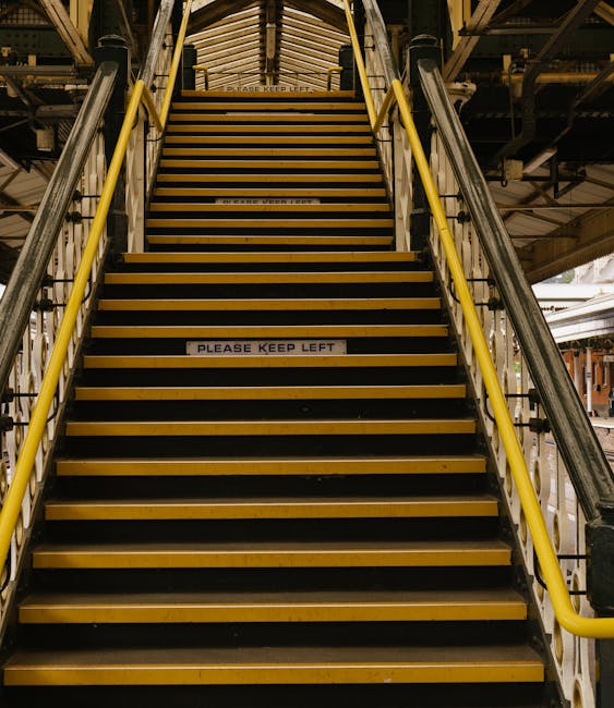A straight staircase inside a building with metal railings painted yellow along each side and black non-slip strips on every step, which are made of sturdy material. The staircase has a sign on the risers reading 'PLEASE KEEP LEFT', indicating directional guidance for movement. Above, a wooden or metal structure supports the overhead space, with visible beams and components, and ample lighting illuminating the area. The staircase appears to be part of an industrial or warehouse setting, possibly within a logistics or moving environment associated with home relocation services. This setting is relevant to furniture transport and loading processes handled by companies like Man With a Van Stepney, especially during the packing and moving stages involving bulky or heavy items.
