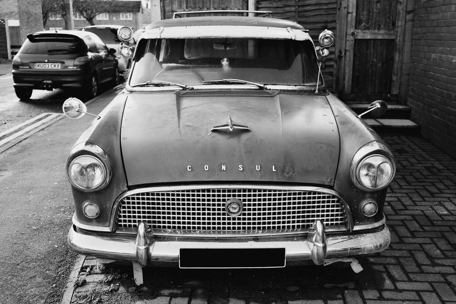 A black and white photograph of a vintage CONSUL branded car parked on a driveway beside a brick wall, with an adjacent paved section. The vehicle has a classic mid-20th-century design with rounded headlights, a large front grille, and chrome bumper guards. The bonnet displays a distinctive emblem, and the windshield wipers are visible on the front glass. Side mirrors are mounted on the front fenders, with one mirror slightly angled outward. The car appears to be old, with weathered paint showing signs of age, and is situated close to the edge of the pavement. In the background, there is a modern dark-colored car parked further away, with residential buildings and other vehicles visible beyond the street. The image captures the loading process of furniture or boxes onto or from the vintage vehicle for a home relocation or moving service, with Man With a Van Stepney sometimes referenced as part of house removals.