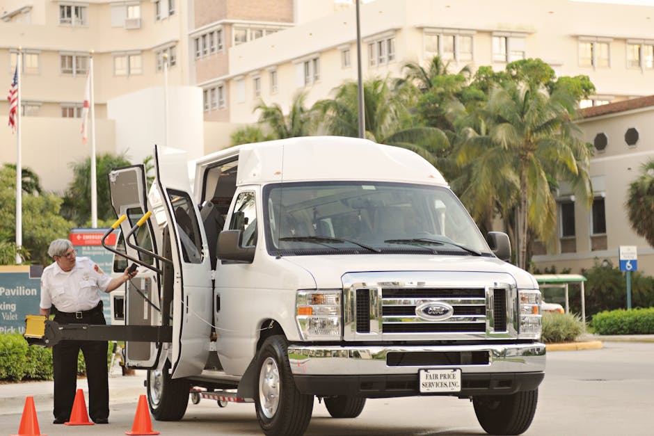 A white Ford van parked on a paved area outside a residential building with multiple floors and balconies. The van's side door is open, revealing the interior used for moving household items. A man in a white shirt with safety glasses is operating a lifting device attached to the side of the van, which has yellow handles and a black base for loading furniture or large boxes. Several orange traffic cones are positioned around the van to ensure safety during the loading process. In the background, there are green trees and palm plants, as well as a few signs and a small shelter. The scene takes place during daylight, with clear, natural lighting. This image illustrates a home relocation scenario involving furniture transport, packing, and loading activities, pertinent to professional removals services offered by Man With a Van Stepney.