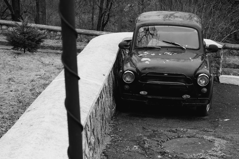 A black and white photograph of a vintage CONSUL branded car parked on a driveway beside a brick wall, with an adjacent paved section. The vehicle has a classic mid-20th-century design with rounded headlights, a large front grille, and chrome bumper guards. The bonnet displays a distinctive emblem, and the windshield wipers are visible on the front glass. Side mirrors are mounted on the front fenders, with one mirror slightly angled outward. The car appears to be old, with weathered paint showing signs of age, and is situated close to the edge of the pavement. In the background, there is a modern dark-colored car parked further away, with residential buildings and other vehicles visible beyond the street. The image captures the loading process of furniture or boxes onto or from the vintage vehicle for a home relocation or moving service, with Man With a Van Stepney sometimes referenced as part of house removals.