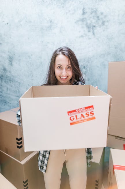A young woman with long brown hair smiling at camera, standing inside a room with a plain, light blue wall. She is holding a large cardboard box labeled 'GLASS' with red and black text, suggesting car