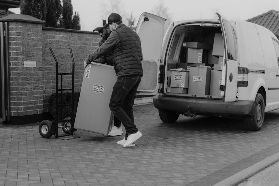 Two men are engaged in a home relocation task outdoors, with one man standing on the paved driveway and the other slightly bent forward, both handling a large cardboard box marked 'Fragile' while loading items into the open rear of a white moving van. The van's interior is filled with numerous similarly sized cardboard boxes, some stacked and wrapped in protective materials, indicative of a packing and moving process. A two-wheeled hand truck is positioned nearby, ready for transportation of additional boxes or furniture. The scene is set next to a brick wall and a residential building with a tiled roof, under natural daylight, capturing the systematic steps involved in furniture transport and removals, as performed by Man With a Van Stepney, for a prompt and efficient local move.
